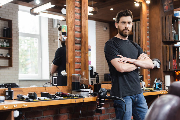 confident hairdresser standing with crossed arms and looking at camera at workplace in barbershop - Photo, Image