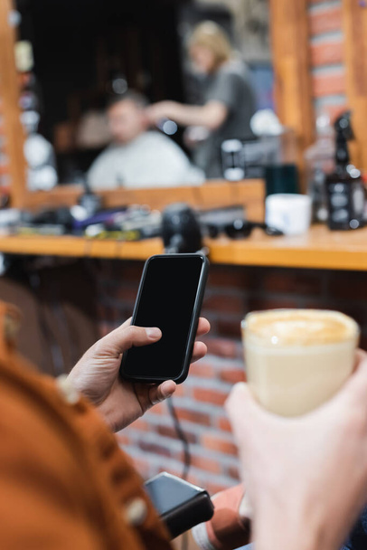 vista recortada del hombre borroso sosteniendo teléfono inteligente con pantalla en blanco y vidrio de capuchino en la barbería - Foto, imagen
