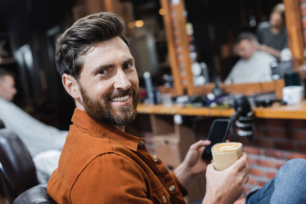homme barbu avec verre de cappuccino et téléphone cellulaire flou souriant à la caméra dans le salon de coiffure - Photo, image