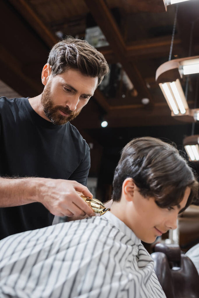 bearded barber trimming neck of teenage boy with hair clipper - Photo, Image