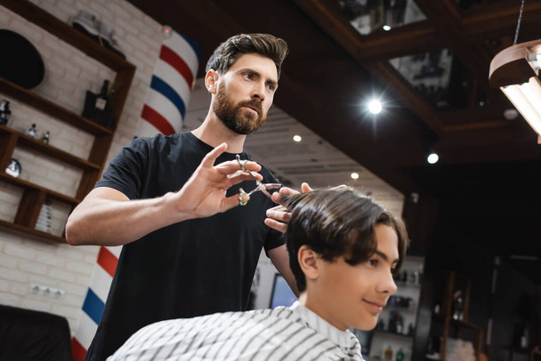 bajo ángulo vista de barbero barbudo haciendo corte de pelo a cliente adolescente en salón de belleza - Foto, Imagen