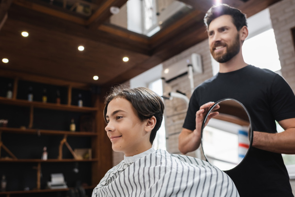 heureux coiffeur tenant miroir près heureux adolescent client dans salon de coiffure - Photo, image