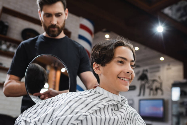 blurred hairstylist holding mirror near happy teenage boy sitting in hairdressing cape - Photo, Image