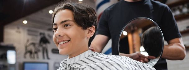 wesoły nastolatek uśmiechnięty obok fryzjera z lustrem w salonie fryzjerskim, sztandar - Zdjęcie, obraz