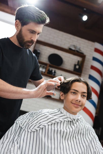 Friseur schneidet Haare eines brünetten Teenagers im Friseurumhang - Foto, Bild