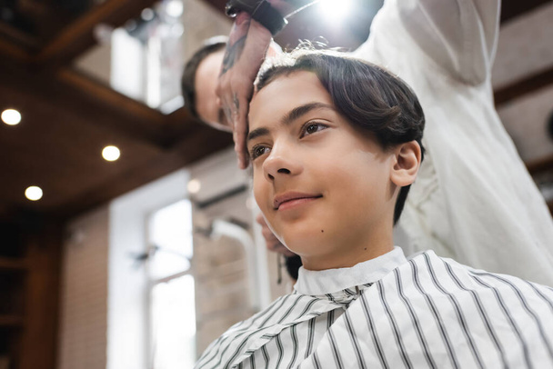 low angle view of smiling teenage guy near blurred hairstylist in barbershop - Zdjęcie, obraz