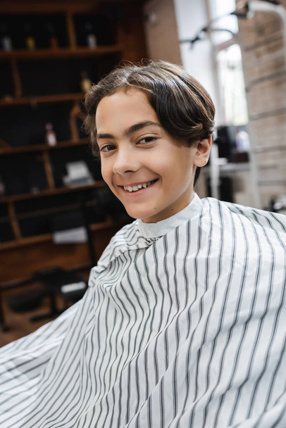 szczęśliwy nastolatek chłopiec w fryzjer peleryna patrząc w aparat fotograficzny w fryzjer - Zdjęcie, obraz