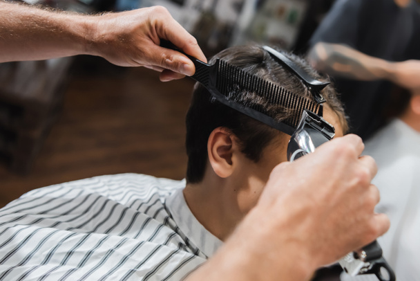 Hairdresser holding comb and clipper while trimming hair of teen boy in beauty salon  - Фото, изображение