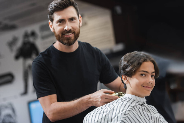 Улыбающийся парикмахер держит триммер рядом с клиентом и смотрит в камеру в салоне красоты  - Фото, изображение