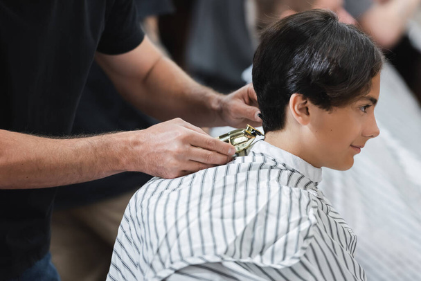 Friseur hält Haarschneidemaschine in der Nähe von Haaren von Teenie-Jungen in Umhang in Schönheitssalon  - Foto, Bild