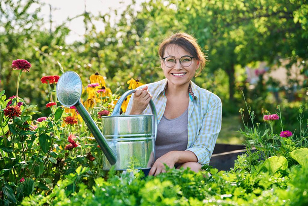 Outdoor portrait woman with watering can in vegetable garden in summer season. Smiling middle aged female looking at camera, beds with vegetables herbs flowers background - Zdjęcie, obraz