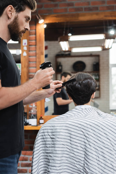 Парикмахер держит распылитель, расчесывая волосы клиента в салоне красоты  - Фото, изображение
