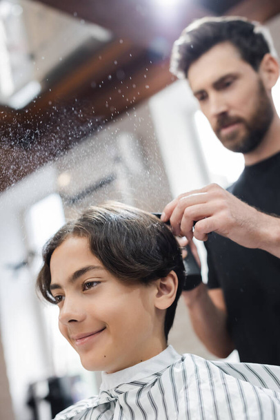 Uśmiechnięty klient siedzi w pobliżu rozmazanego fryzjera z opryskiwaczem w salonie piękności  - Zdjęcie, obraz
