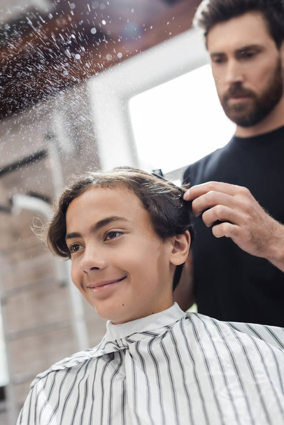 Cheerful teenager in cape sitting near blurred hairdresser spraying water in beauty salon  - Фото, зображення