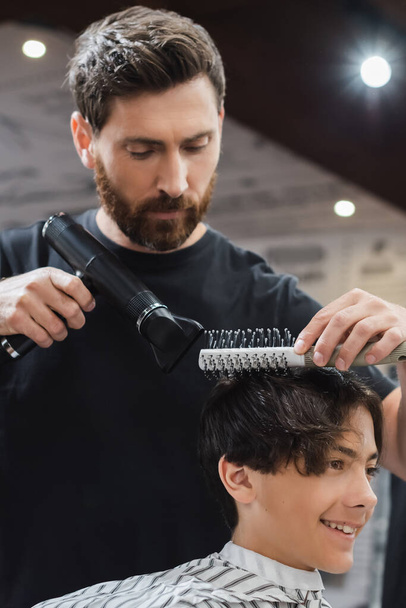 Barber drying and combing hair of smiling teen boy in beauty salon  - 写真・画像