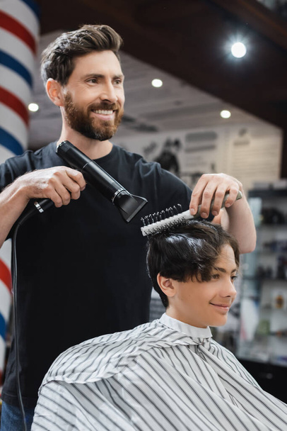 Glücklicher Friseur trocknet die Haare lächelnder Teenager im Friseurumhang im Schönheitssalon  - Foto, Bild
