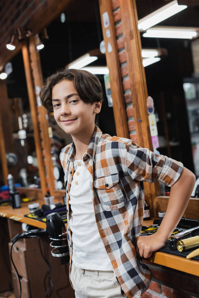 Positive teenage boy looking at camera near worktop and mirror in barbershop  - Fotó, kép