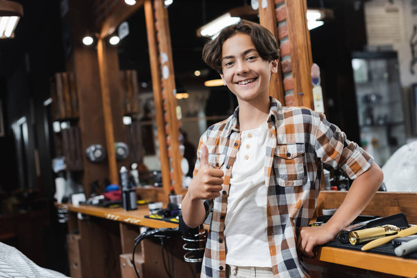 Vrolijke tiener jongen tonen duim omhoog in de buurt kappersgereedschap en spiegel in schoonheidssalon  - Foto, afbeelding