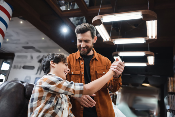 Cheerful barber holding hand of teenage client in barbershop  - Фото, изображение