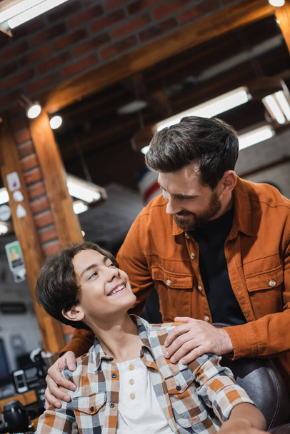 Smiling barber hugging teenager in armchair in barbershop  - 写真・画像