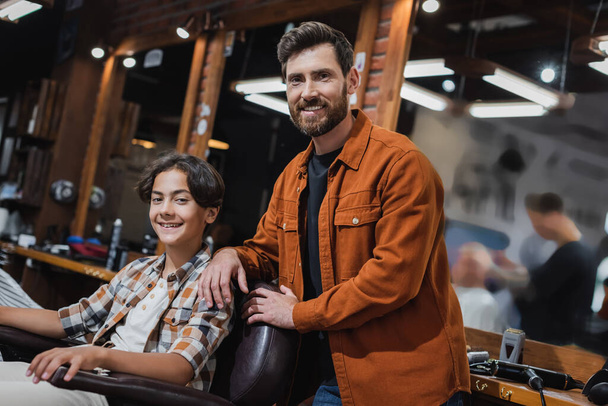 Позитивный бородатый парикмахер улыбается в камеру рядом с клиентом-подростком в парикмахерской  - Фото, изображение