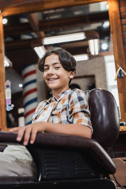 Teenage client smiling at camera while sitting in armchair in barbershop  - Foto, afbeelding