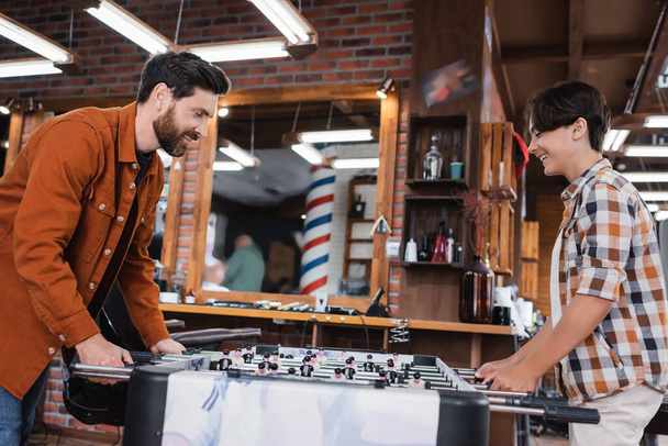 Side view of smiling father and son playing table football in barbershop  - Foto, Imagem