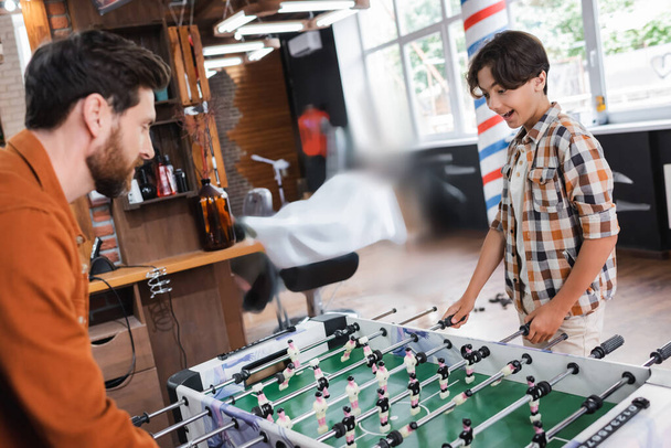 Excited teenager playing table football with blurred father in barbershop  - Fotografie, Obrázek