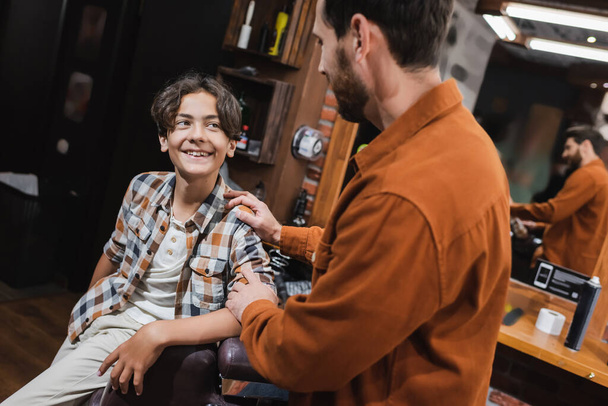 Adolescent joyeux regardant barbier flou dans le salon de coiffure  - Photo, image