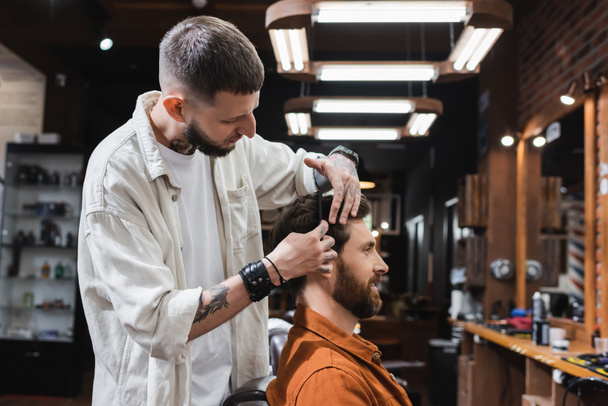 Tätowierter Friseur kämmt Haare eines lächelnden Mannes im Friseursalon  - Foto, Bild