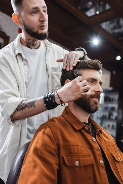 Tatuado barbeiro pentear o cabelo do homem satisfeito na barbearia  - Foto, Imagem