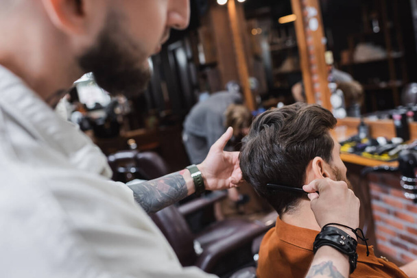 Blurred tattooed barber combing hair of man in barbershop  - Φωτογραφία, εικόνα