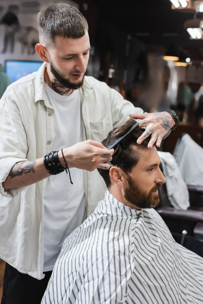 Tattooed barber combing hair of bearded customer in barbershop  - Photo, Image