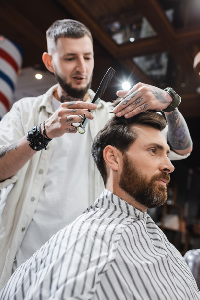 Низький кут зору розмитого перукаря, що тримає ножиці і гребінець, торкаючись волосся клієнта в перукарні
  - Фото, зображення