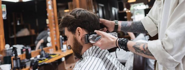 Peluquero tatuado que sostiene trimmer cerca de cliente barbudo en barbería, bandera  - Foto, imagen
