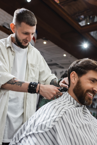 Sonriente peluquero tatuado recorte cuello de cliente barbudo en la barbería  - Foto, Imagen