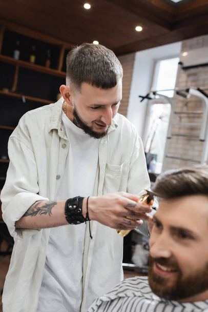 Positive barber holding hair clipper near blurred bearded client in barbershop  - Фото, изображение