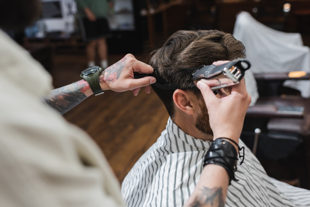 Tattooed hairstylist trimming and combing hair of customer in barbershop  - 写真・画像