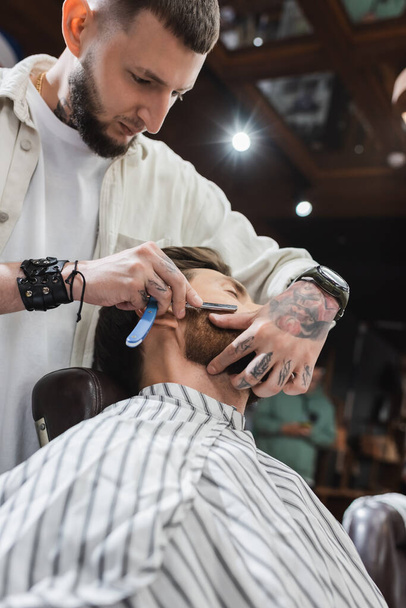 низкий угол зрения татуированный парикмахер бритья бороду человека с прямой бритвой - Фото, изображение