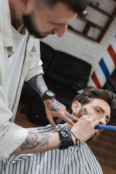 blurred hairdresser in leather bracelet shaving cheek of man with straight leather - Foto, Bild