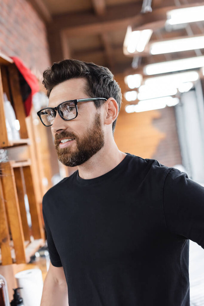 Bearded barber in eyeglasses and black t-shirt standing in barbershop  - Zdjęcie, obraz