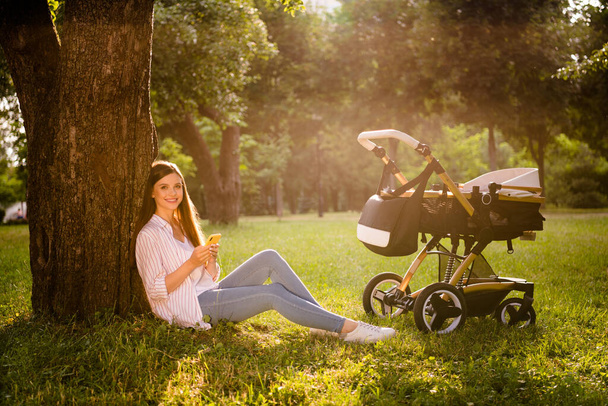 Full length photo of cute mom sit lawn urban street park use gadget blog while infant kid sleep pram outdoors. - Foto, Bild