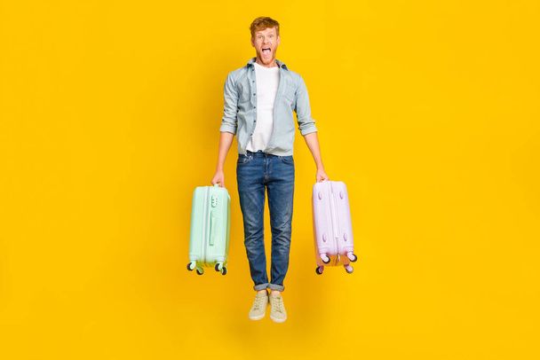 Full length photo of impressed handsome guy red hairstyle wear blue shirt jeans jump scream hold bags isolated on yellow color background. - Zdjęcie, obraz