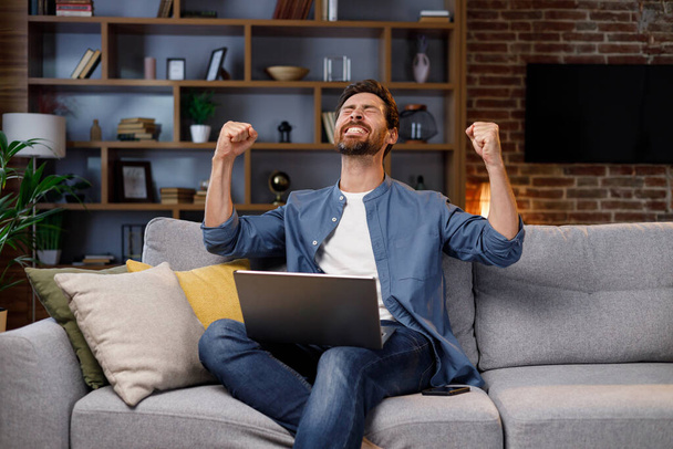 Man sitting on couch working on laptop, receiving bank loan approval notification email, feeling successful, clenching fists, making yes gesture. Sincere emotions, victory result, achievement concept. - Fotoğraf, Görsel