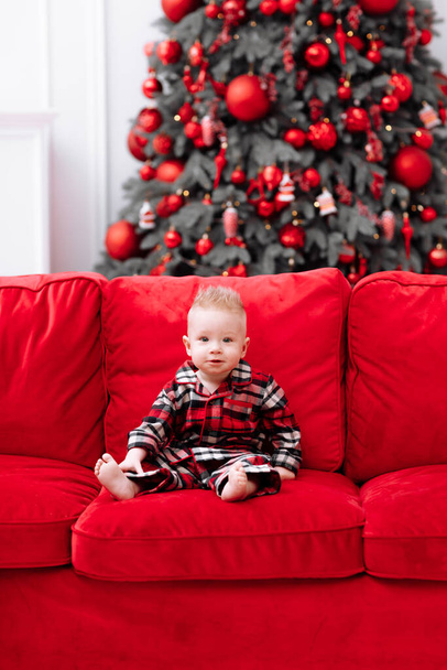adorable little baby boy in a red christmas outfit sitting on red sofa in front of a christmas tree. Beautiful little baby boy celebrating Christmas.Family on Christmas eve at fireplace - Fotó, kép