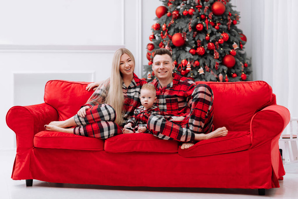 The theme of the family holiday is New year and Christmas. Young European family in the same pyjamas: mom, dad, baby boy are sitting on the red sofa by the festive Christmas tree on Christmas evening. - 写真・画像