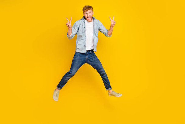 Full length photo of impressed handsome guy blue shirt jeans sneakers flying show v-sign scream yeah isolated on yellow color background. - Фото, зображення