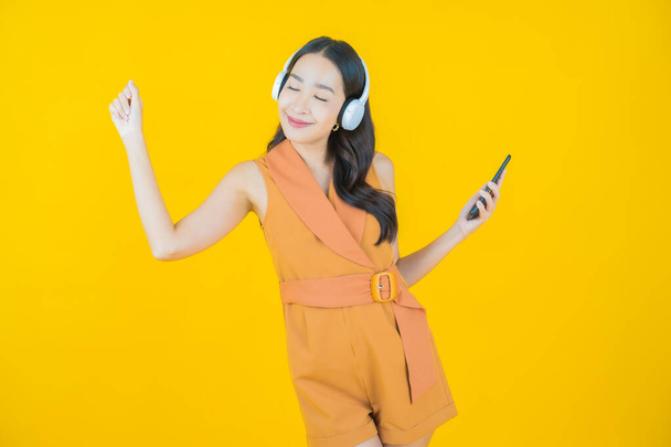 Portrét krásná mladá asijská žena se sluchátkem a chytrý telefon pro poslech hudby na žlutém pozadí - Fotografie, Obrázek