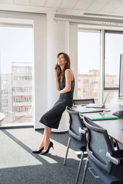 Full length of an attractive businesswoman sitting in office and talking on the phone. Executive female wearing black dress.  - Photo, image