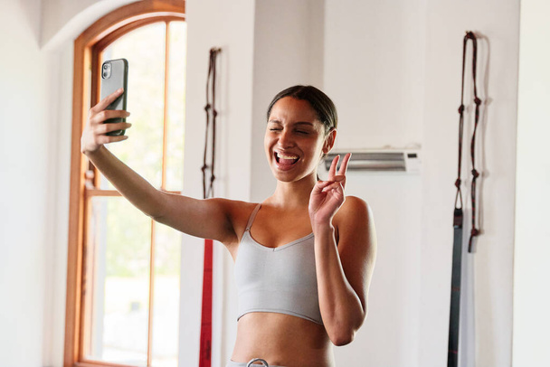 Νεαρή γυναίκα με αυτοπεποίθηση στα αθλητικά να βγάζει selfie με κινητό τηλέφωνο στο γυμναστήριο - Φωτογραφία, εικόνα